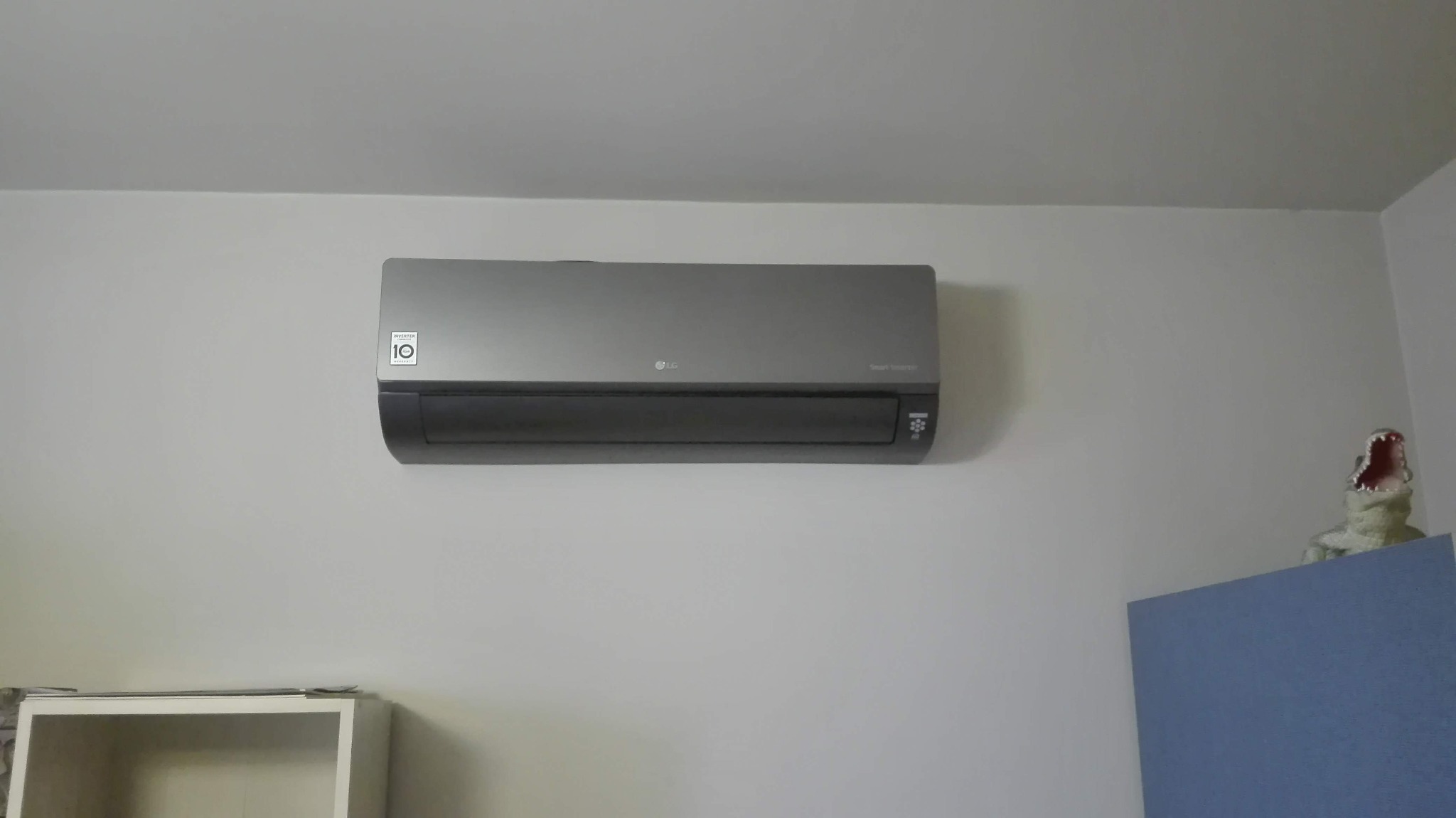Szary klimatyzator LG zamontowany na białej ścianie w pomieszczeniu, z zabawką krokodyla w tle, widać logo 'Inverter 10'.
