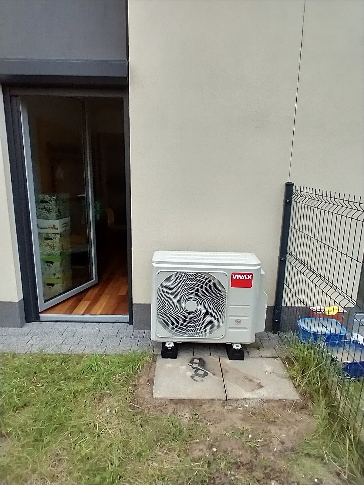 Zewnętrzna jednostka klimatyzacyjna marki Vivax umieszczona na betonowej podstawie obok ściany budynku i otwartego okna tarasowego z widocznym wnętrzem, przylegająca do trawnika i ogrodzenia...