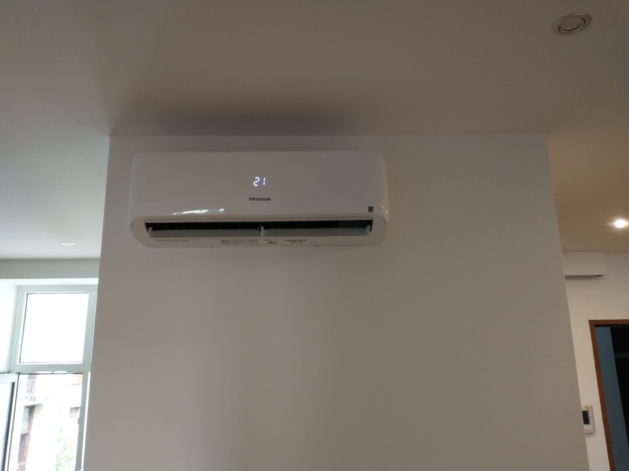 Biały klimatyzator Hisense zamontowany na białej ścianie, wyświetlający temperaturę 21 stopni Celsjusza, widok z dołu w jasnym pomieszczeniu z oknem.