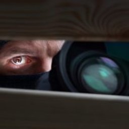 Oczy mężczyzny w czarnej kominiarce, spoglądające przez szparę w drewnianej konstrukcji, z widocznym obiektywem aparatu. Ujęcie z ukrycia.