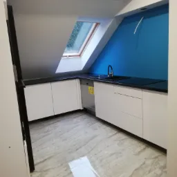 Nowoczesna kuchnia na poddaszu z białymi szafkami, czarnym blatem i niebieską ścianą, oświetlona przez okno dachowe.