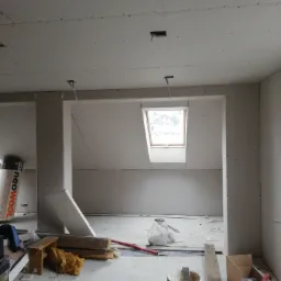 Montaż płyt kartonowo-gipsowych na poddaszu z oknem dachowym, widoczne profile metalowe, puszka po masie szpachlowej Atlas i inne materiały budowlane na podłodze.