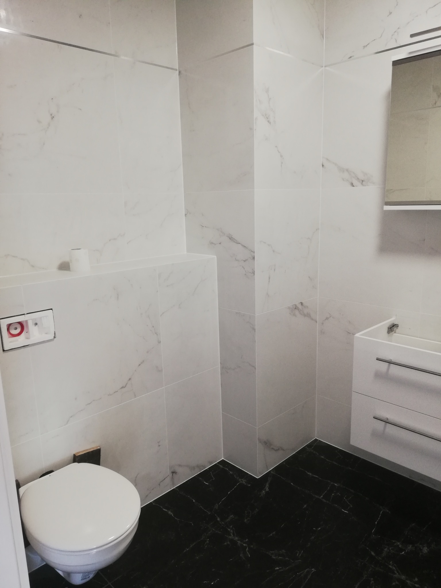 Nowoczesna łazienka z białymi płytkami imitującymi marmur na ścianach, czarną podłogą z wzorem marmuru, podwieszaną toaletą i białą szafką z umywalką.