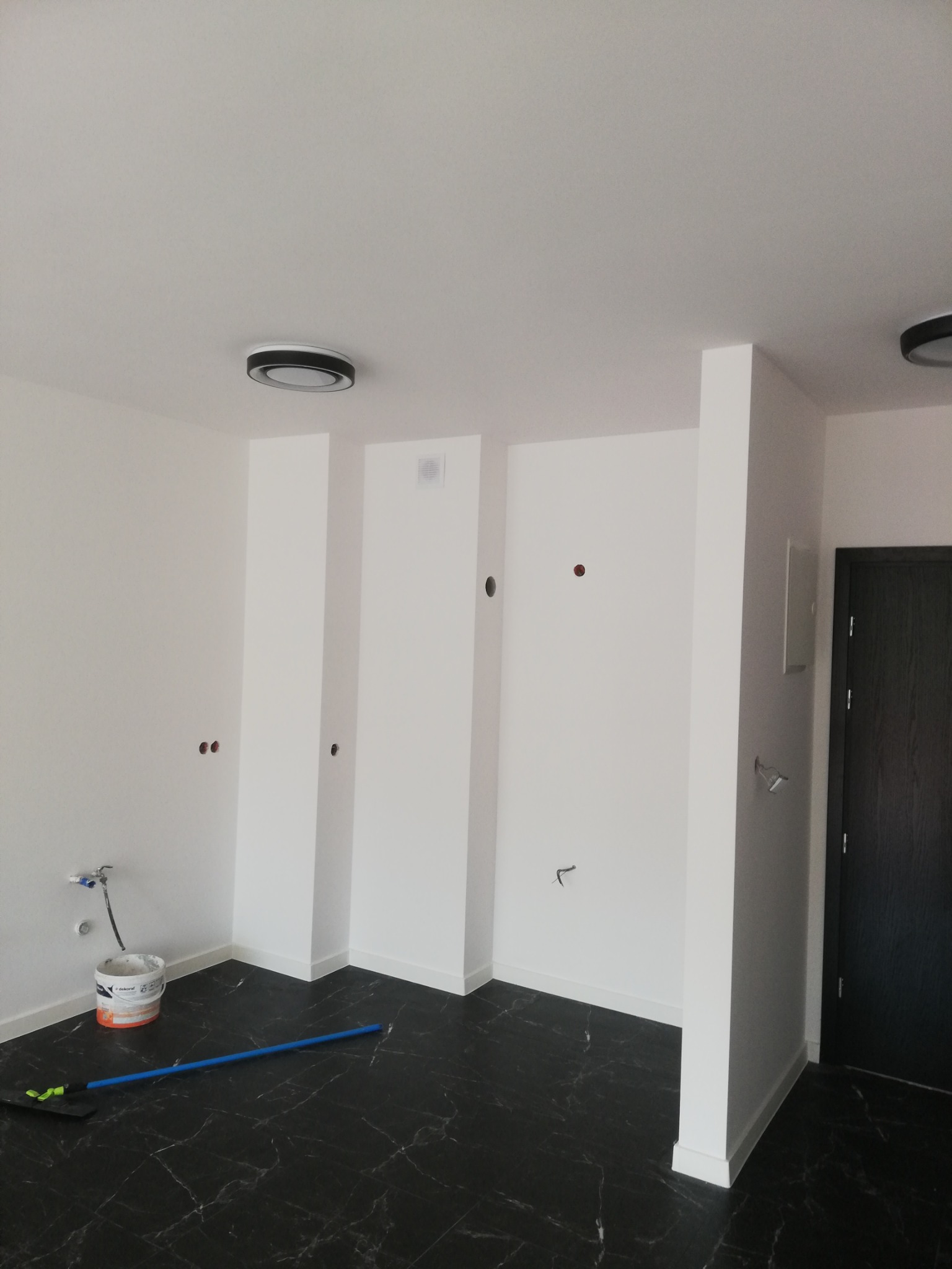 Wnętrze po malowaniu, z czarną marmurową podłogą, białe ściany z widocznymi puszkami instalacyjnymi, wiadro z farbą i mop w rogu.