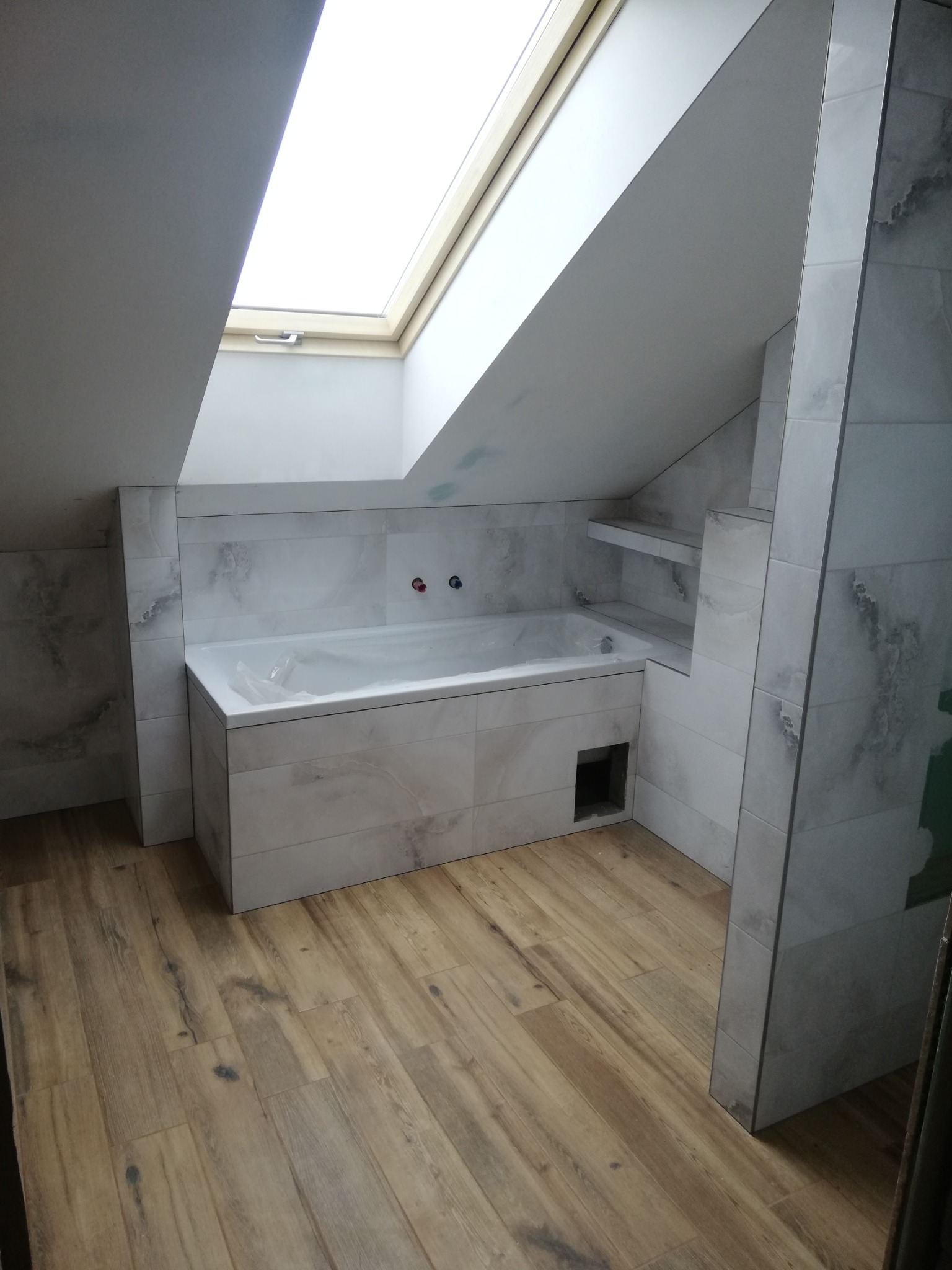 Łazienka na poddaszu z oknem dachowym, wykończona płytkami imitującymi marmur, z zabudowaną wanną i drewnopodobną podłogą.