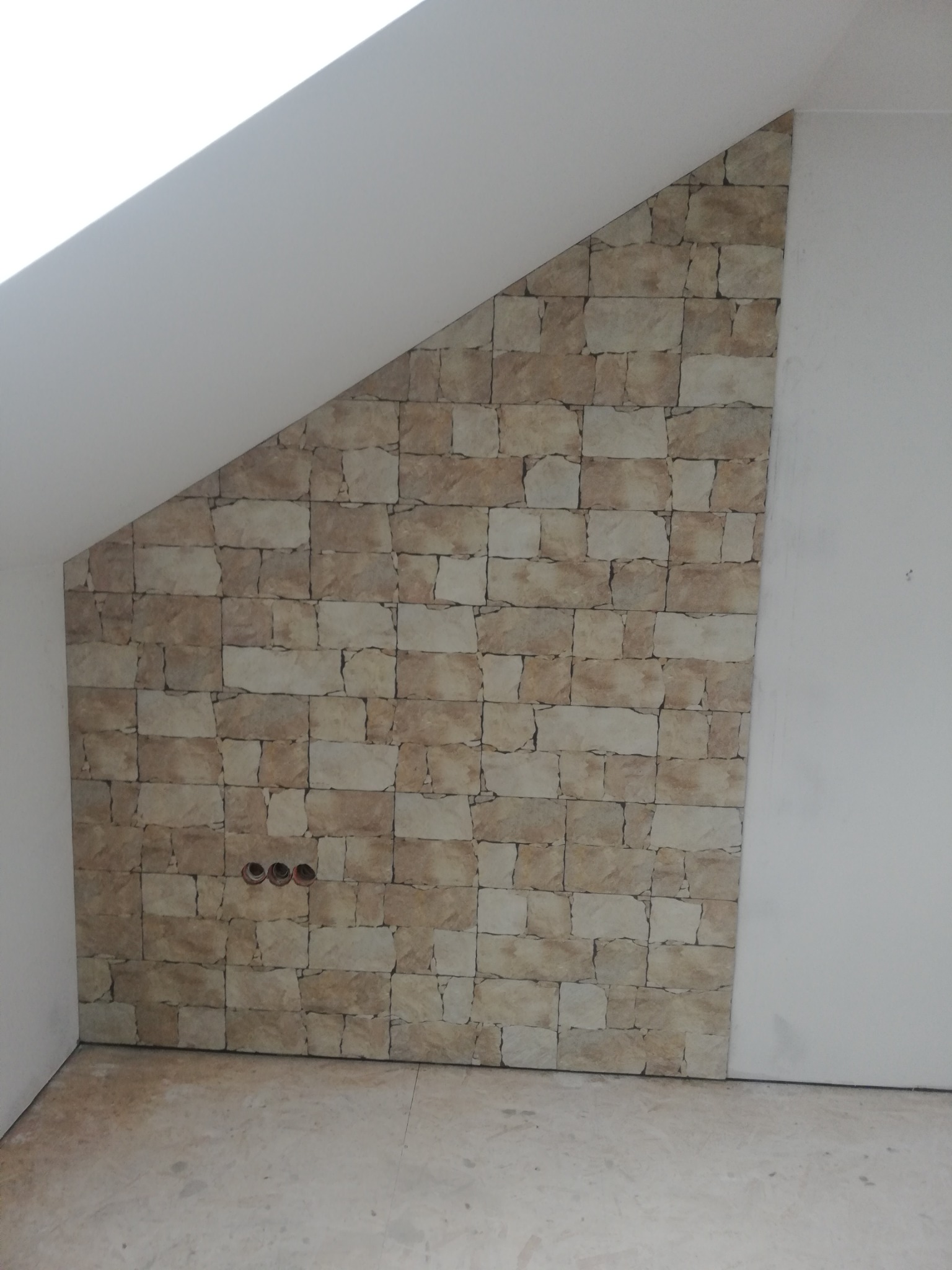 Ściana pod skosem wykończona tapetą imitującą kamień, widoczne trzy otwory na gniazdka elektryczne, surowy stan podłogi i fragment ściany pomalowanej na biało.