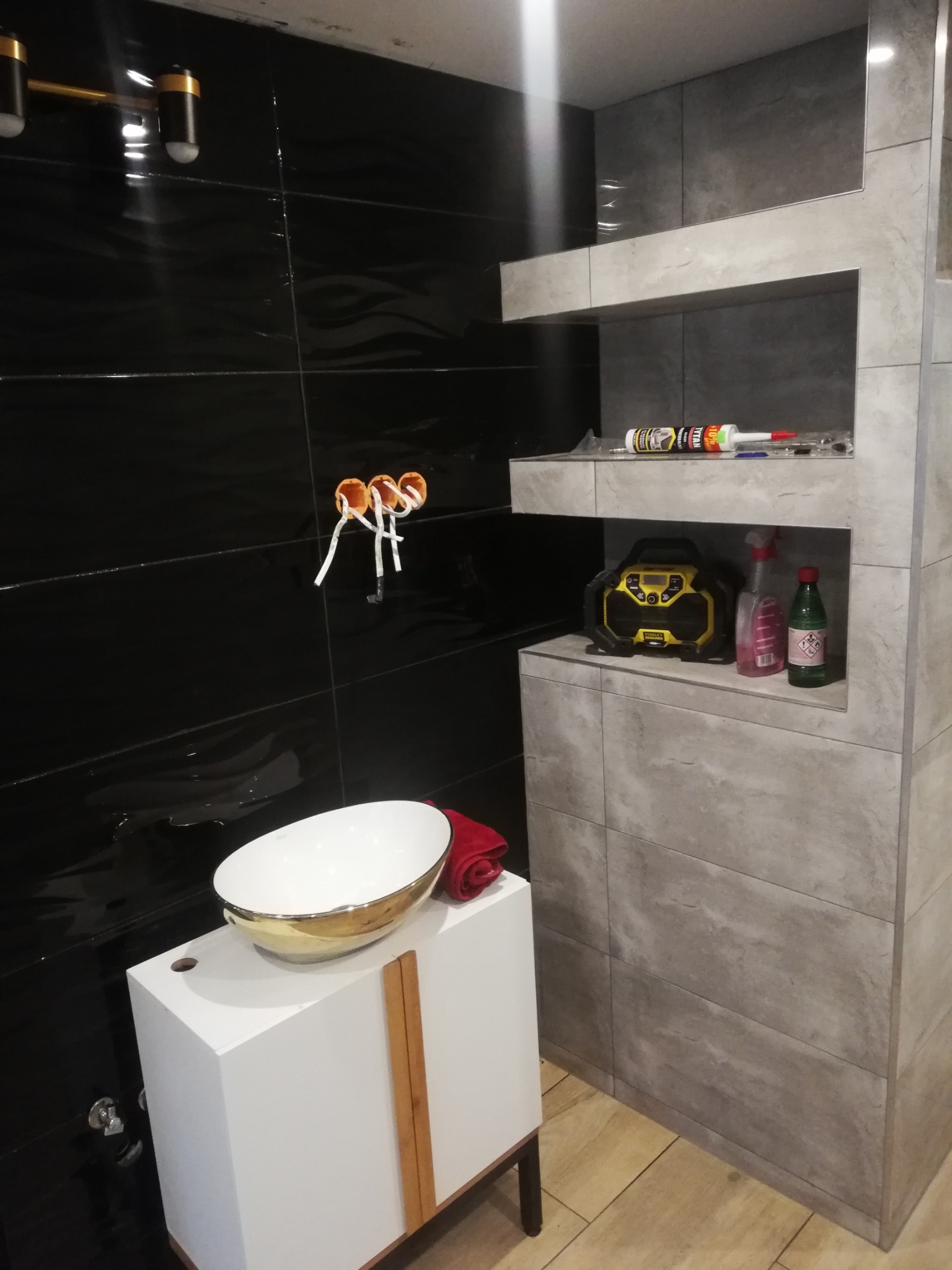 Łazienka w trakcie wykańczania: czarne, błyszczące płytki na ścianie, biała umywalka z elementami złota na szafce, szare płytki imitujące kamień na ścianie z półkami, widoczne puszki elektryczne...