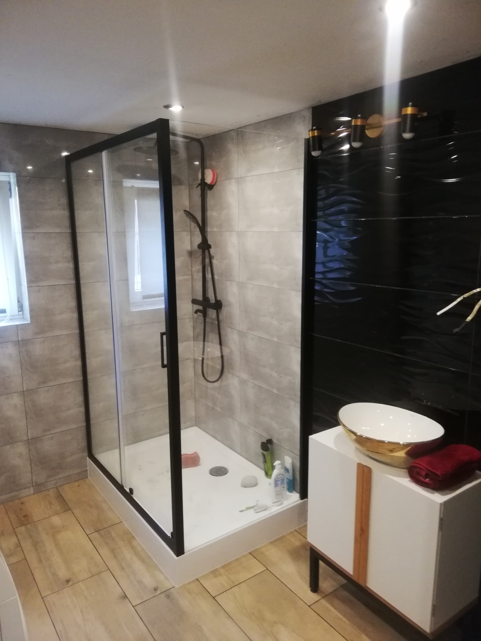 Nowoczesna łazienka z kabiną prysznicową typu walk-in z czarnymi profilami, umywalką nablatową ze złotym akcentem i czarną ścianą z dekoracyjną fakturą fal.