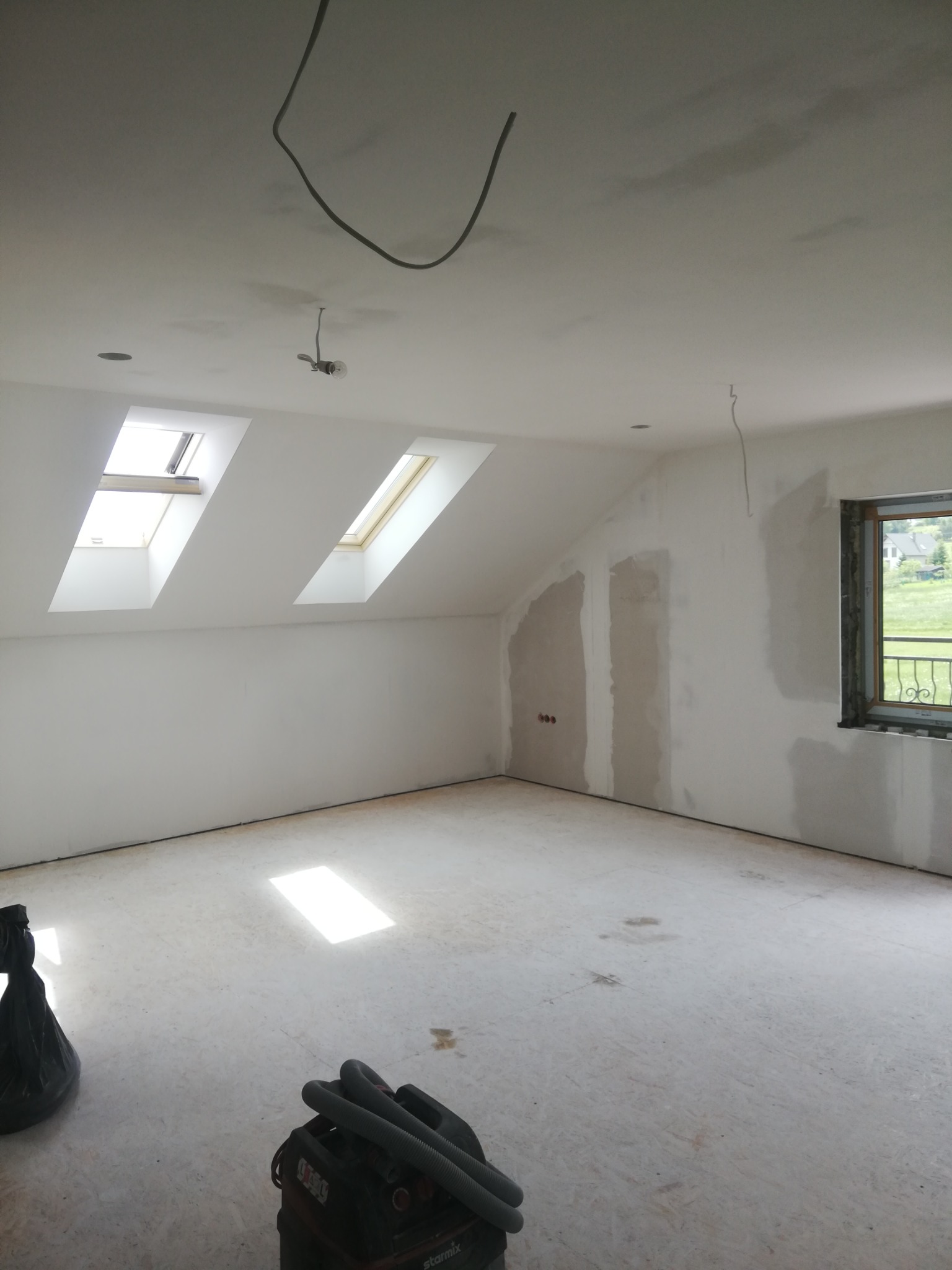 Poddasze w trakcie prac wykończeniowych, widoczne dwa okna dachowe, fragment ściany przygotowanej do szpachlowania i odkurzacz przemysłowy na podłodze z płyt OSB.