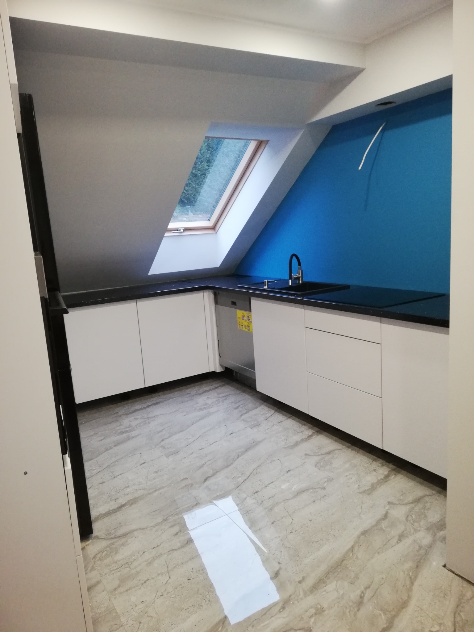 Nowoczesna kuchnia na poddaszu z białymi szafkami, czarnym blatem i niebieską ścianą, oświetlona przez okno dachowe.