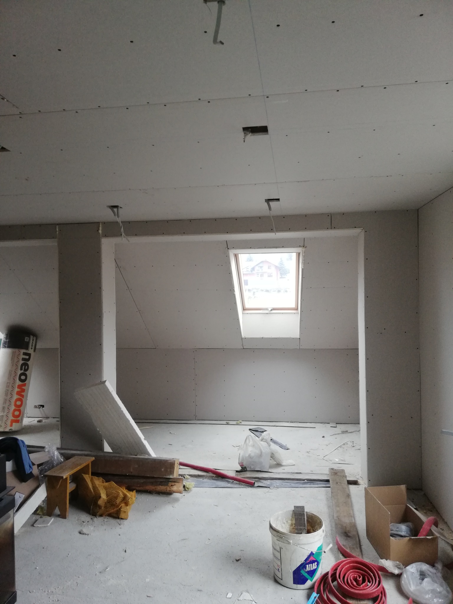 Montaż płyt kartonowo-gipsowych na poddaszu z oknem dachowym, widoczne profile metalowe, puszka po masie szpachlowej Atlas i inne materiały budowlane na podłodze.