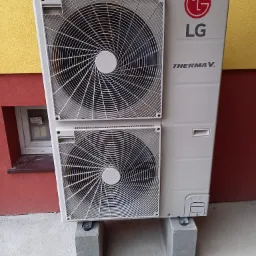 Zewnętrzna jednostka pompy ciepła LG Therma V, umieszczona na betonowych bloczkach przy żółto-czerwonej ścianie budynku z małym okienkiem piwnicznym.