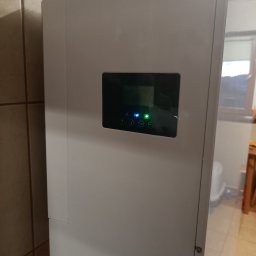 MARGO SOLAR ENERGY - MARIAN MAZUR - Biała obudowa magazynu energii z czarnym panelem sterowania z ikonami i zieloną diodą LED, widoczna w rogu pomieszczenia z brązowymi płytkami.