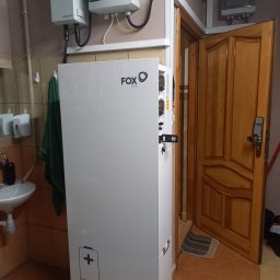 MARGO SOLAR ENERGY - MARIAN MAZUR - Biały moduł magazynu energii Fox ESS z ikoną baterii w pomieszczeniu z umywalką i otwartymi drzwiami.