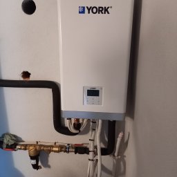 MARGO SOLAR ENERGY - MARIAN MAZUR - Biała jednostka grzewcza marki York zamontowana na szarej ścianie, widoczne rury, zawory i filtry, ściana z niedoskonałościami i śladami wiercenia.