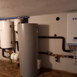 MARGO SOLAR ENERGY - MARIAN MAZUR - Instalacja grzewcza w piwnicy z dwoma białymi zbiornikami, szarym zbiornikiem Ferroli i wiszącym białym urządzeniem York z czarnymi rurami, widoczne zawory i osprzęt hydrauliczny.
