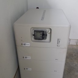 MARGO SOLAR ENERGY - MARIAN MAZUR - Biały, modułowy magazyn energii Fox ESS z widocznym wyłącznikiem DC, umieszczony w pomieszczeniu w Raba Wyżna, z widocznymi naklejkami z kodami QR i regulowanymi nóżkami.