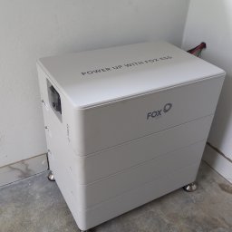 MARGO SOLAR ENERGY - MARIAN MAZUR - Biały, modułowy magazyn energii Fox ESS na srebrnych nóżkach, ustawiony w rogu pomieszczenia z betonową podłogą i białymi ścianami, widoczne kable wychodzące ze ściany.