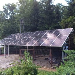 MARGO SOLAR ENERGY - MARIAN MAZUR - Instalacja fotowoltaiczna na wiacie samochodowej z widocznym inwerterem, otoczona zielenią i drzewami, w tle słup energetyczny.