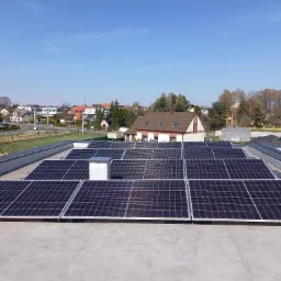 Rząd paneli słonecznych na płaskim dachu budynku, widok z g&oacute;ry w słoneczny dzień, w tle zabudowa jednorodzinna.