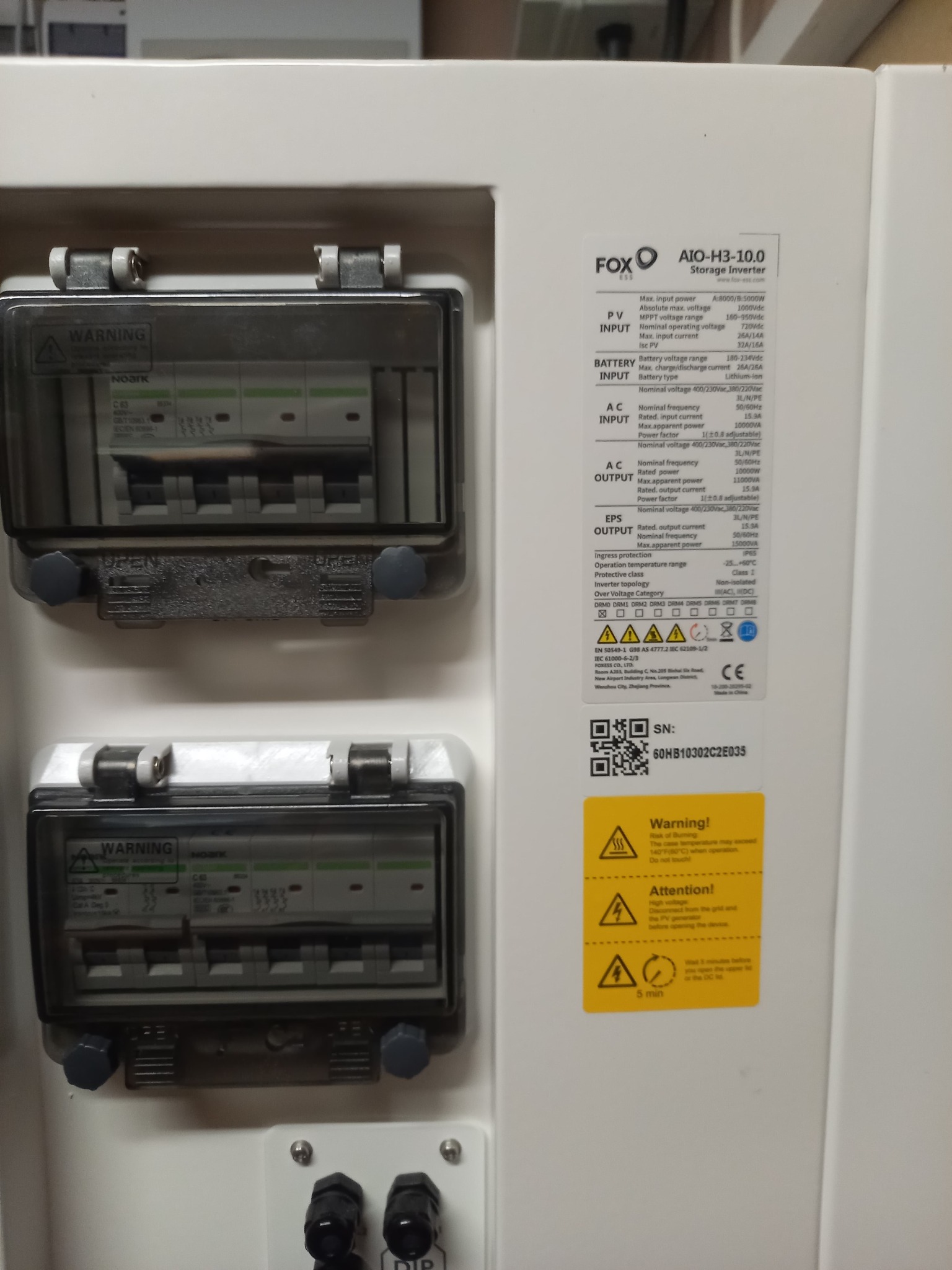 Szczegółowe ujęcie białej obudowy inwertera FOX AIO-H3-10.0 z widocznymi zabezpieczeniami, specyfikacją techniczną, kodem QR, ostrzeżeniami i oznaczeniami certyfikacyjnymi.