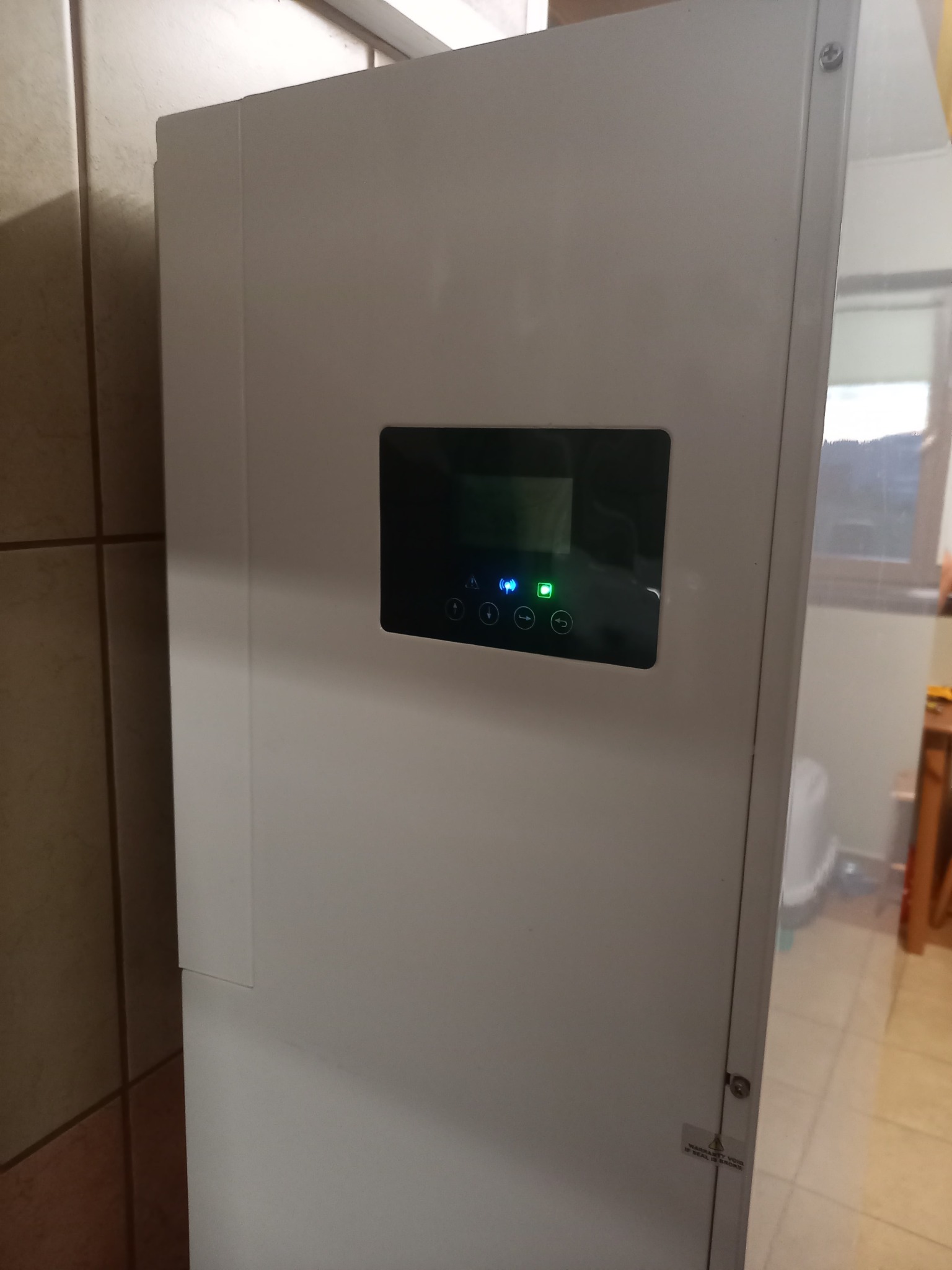 Biała obudowa magazynu energii z czarnym panelem sterowania z ikonami i zieloną diodą LED, widoczna w rogu pomieszczenia z brązowymi płytkami.