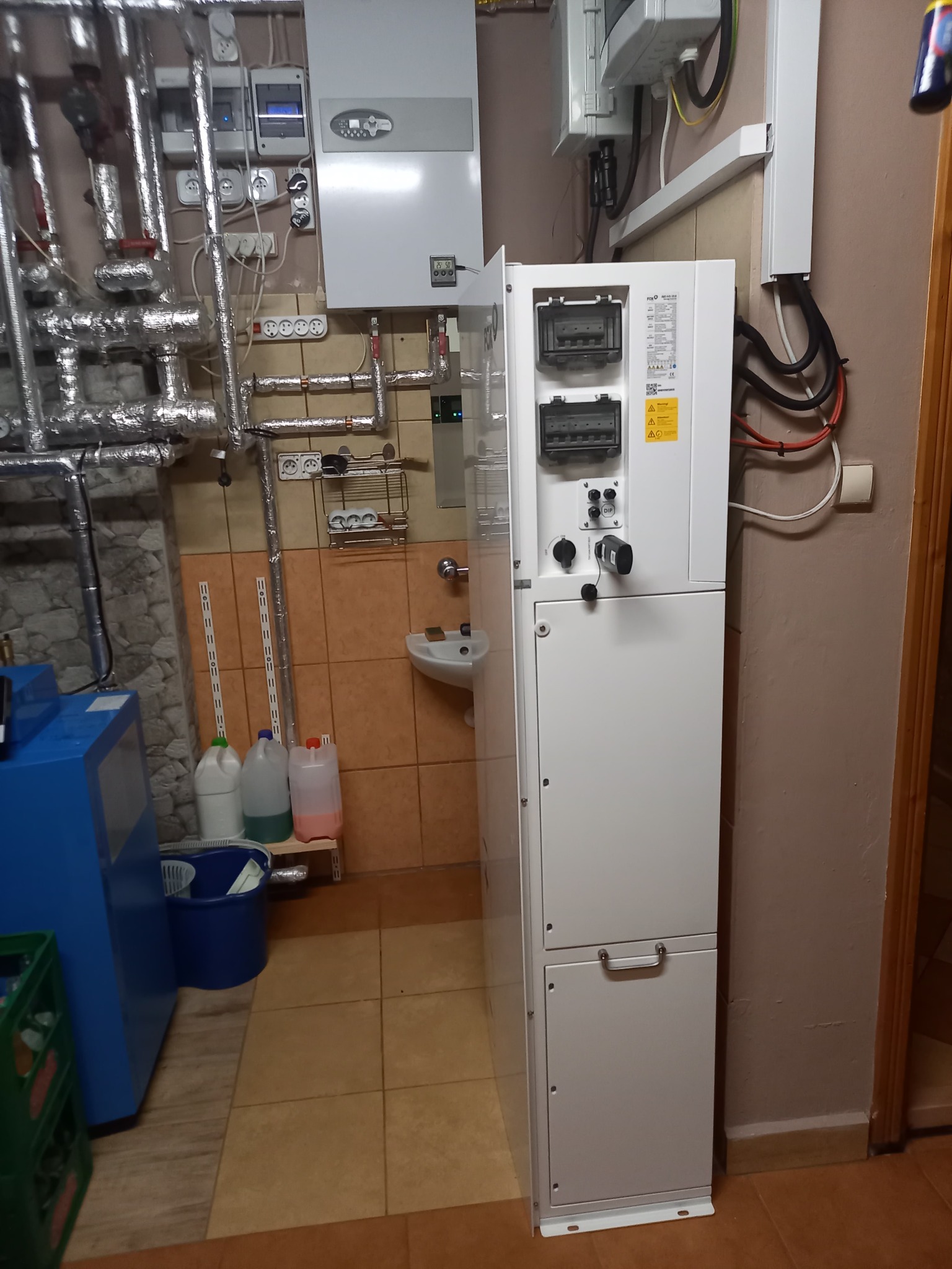 Biała, pionowa pompa ciepła w kotłowni z widocznymi rurami, instalacją elektryczną i umywalką w tle.