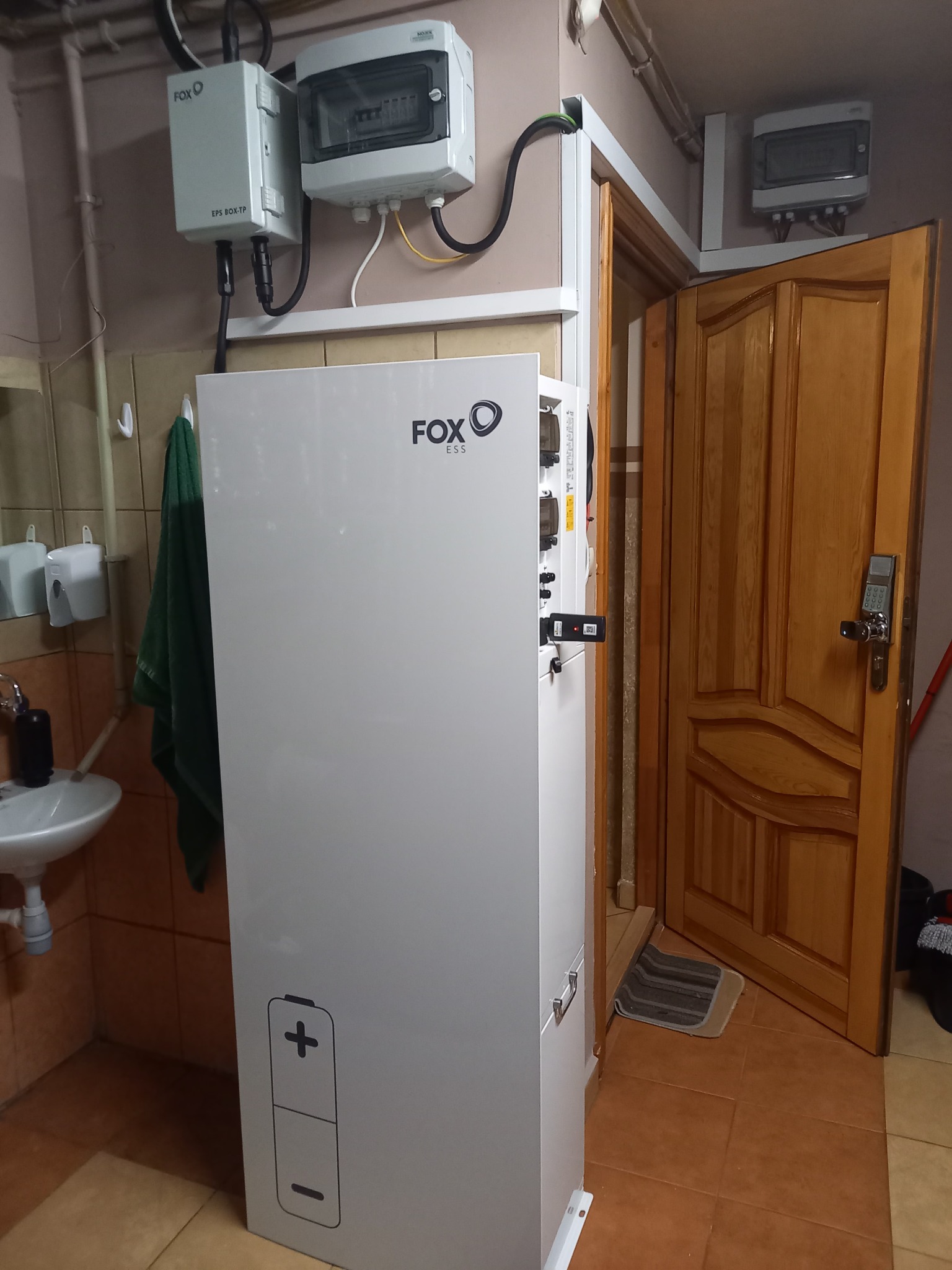 Biały moduł magazynu energii Fox ESS z ikoną baterii w pomieszczeniu z umywalką i otwartymi drzwiami.