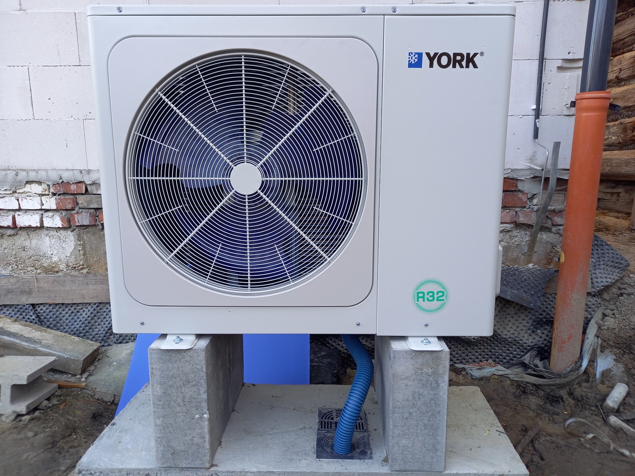 Zewnętrzna jednostka klimatyzacyjna marki York, zamontowana na betonowych bloczkach, z widoczną niebieską rurą odprowadzającą skropliny, na tle ściany z białego bloczka i fragmentu drewnianej...