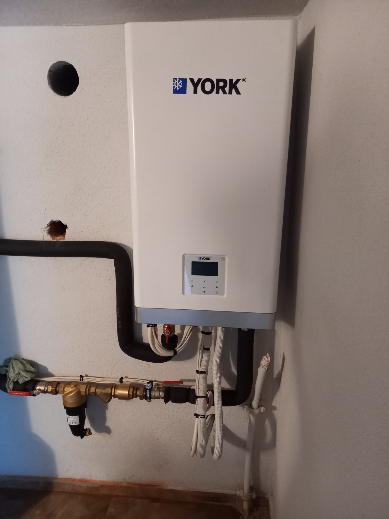 Biała jednostka grzewcza marki York zamontowana na szarej ścianie, widoczne rury, zawory i filtry, ściana z niedoskonałościami i śladami wiercenia.