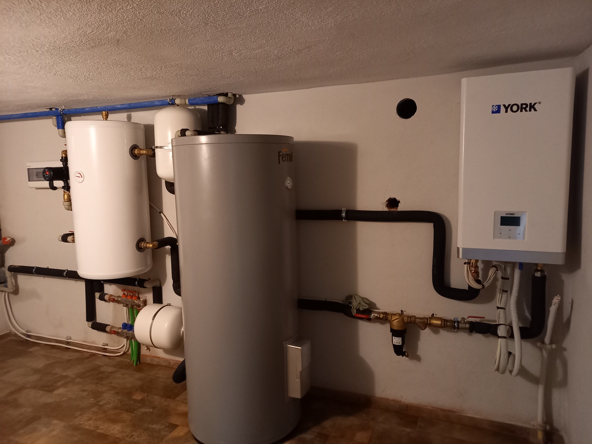 Instalacja grzewcza w piwnicy z dwoma białymi zbiornikami, szarym zbiornikiem Ferroli i wiszącym białym urządzeniem York z czarnymi rurami, widoczne zawory i osprzęt hydrauliczny.