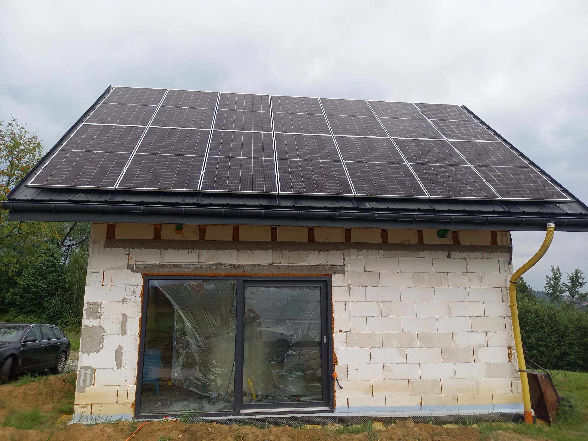 Dom w trakcie budowy z panelami słonecznymi na dachu, okna zasłonięte folią ochronną, widoczny fragment samochodu osobowego.