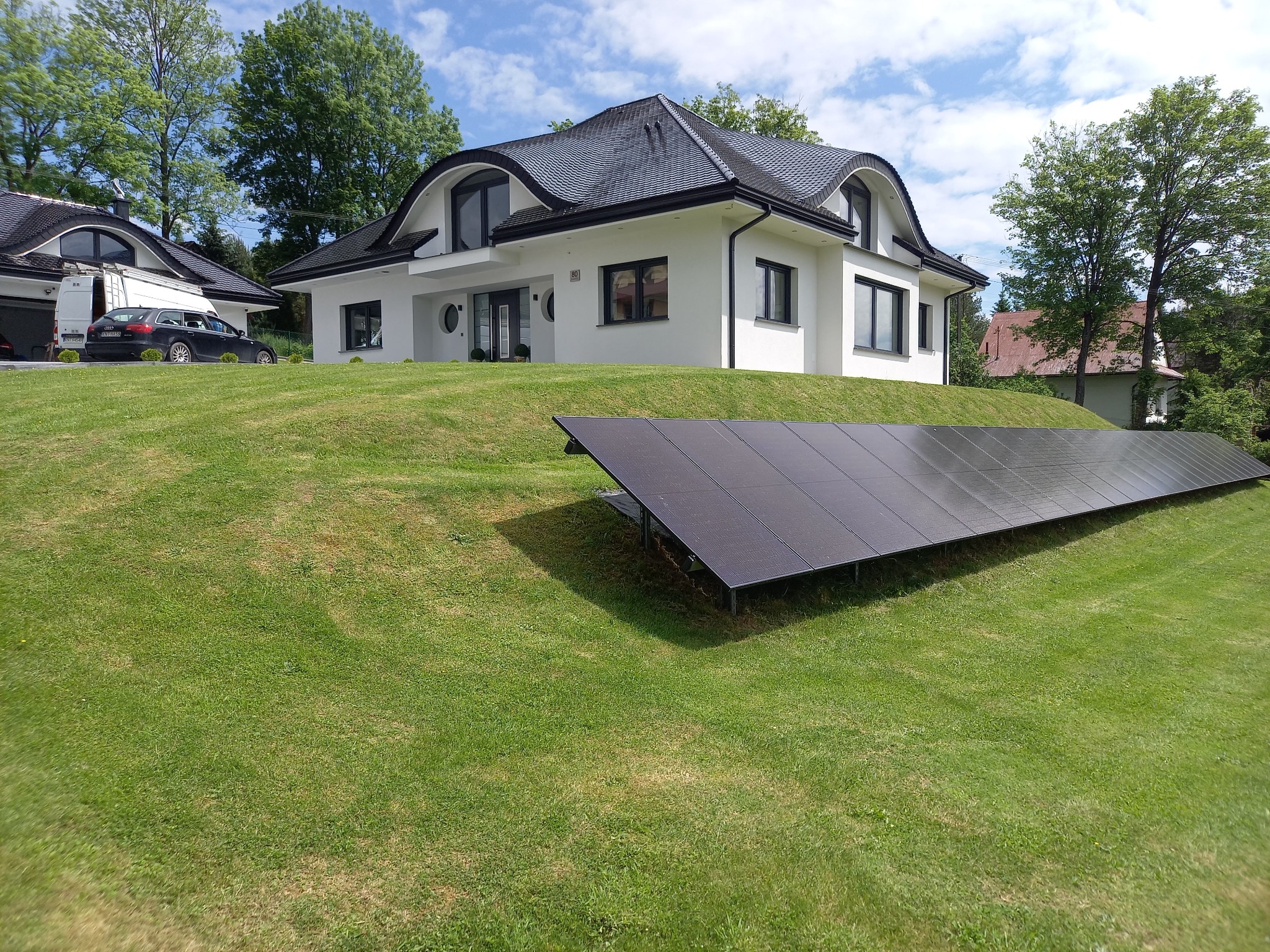 Nowoczesny dom jednorodzinny z zainstalowanymi panelami słonecznymi na trawiastym zboczu przed budynkiem, widoczny biały van w tle.