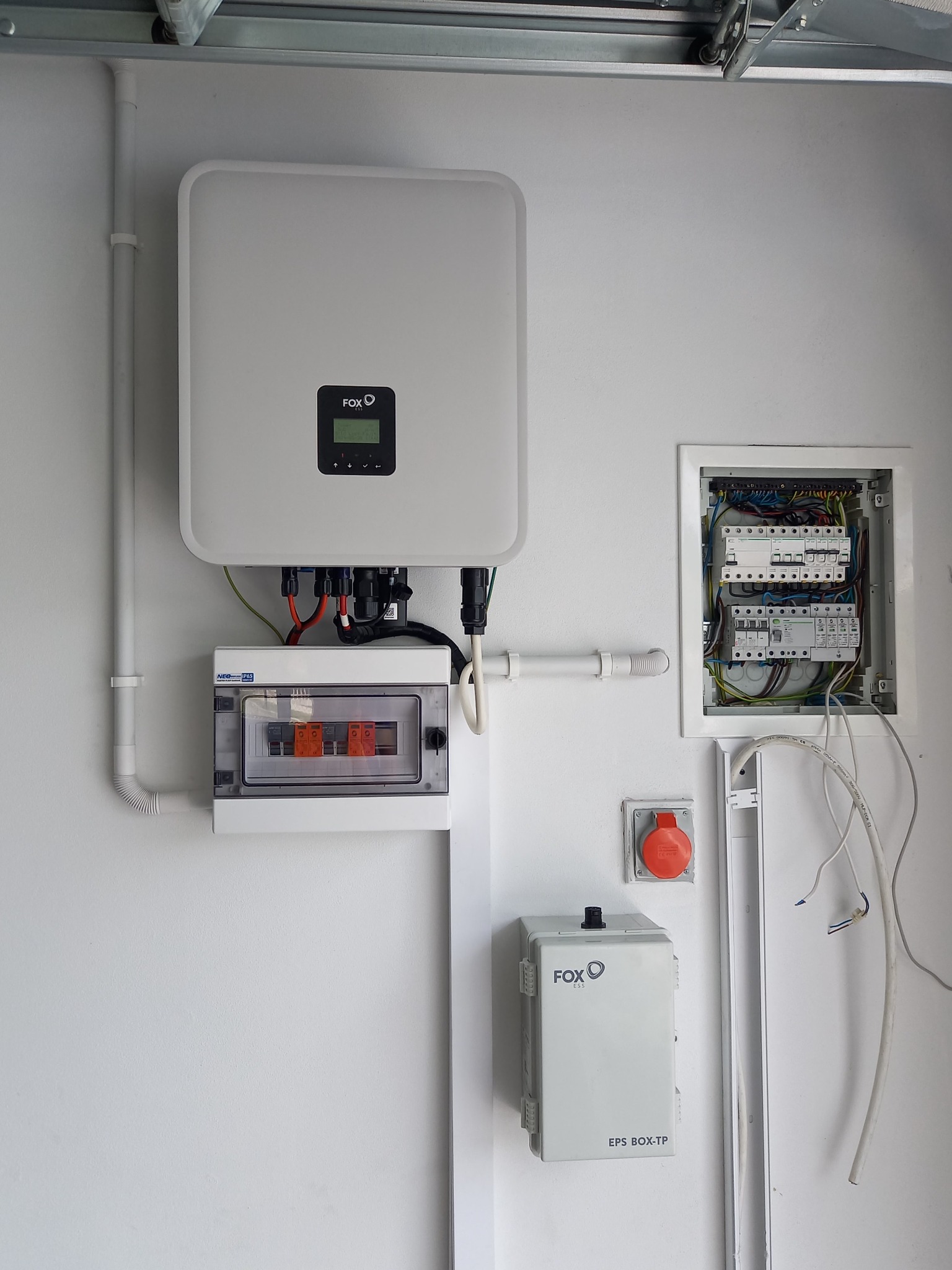Biały falownik z wyświetlaczem marki FOX, pod nim skrzynka z bezpiecznikami, obok skrzynka z okablowaniem i gniazdo siłowe na białej ścianie wewnątrz budynku.