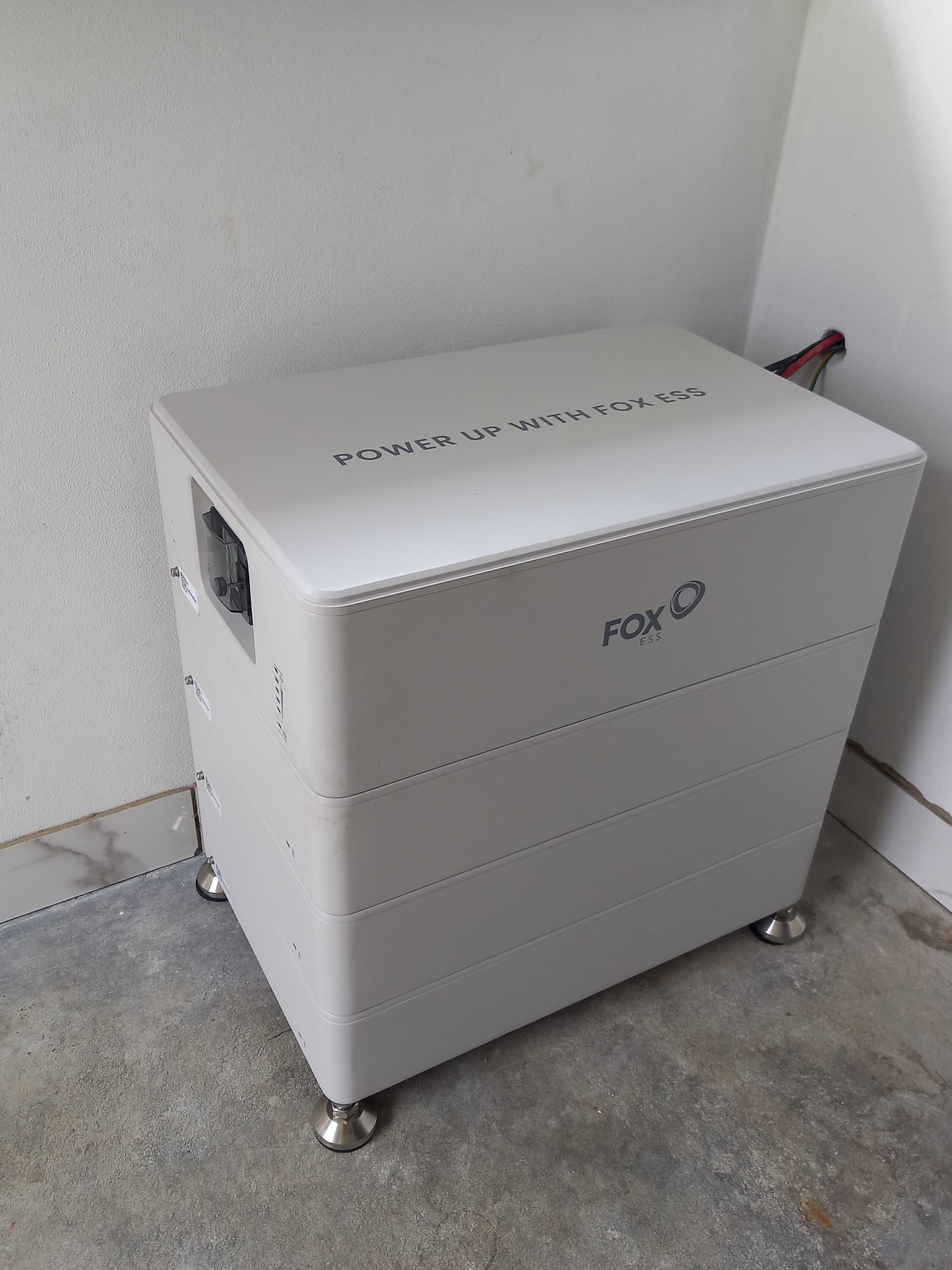 Biały, modułowy magazyn energii Fox ESS na srebrnych nóżkach, ustawiony w rogu pomieszczenia z betonową podłogą i białymi ścianami, widoczne kable wychodzące ze ściany.