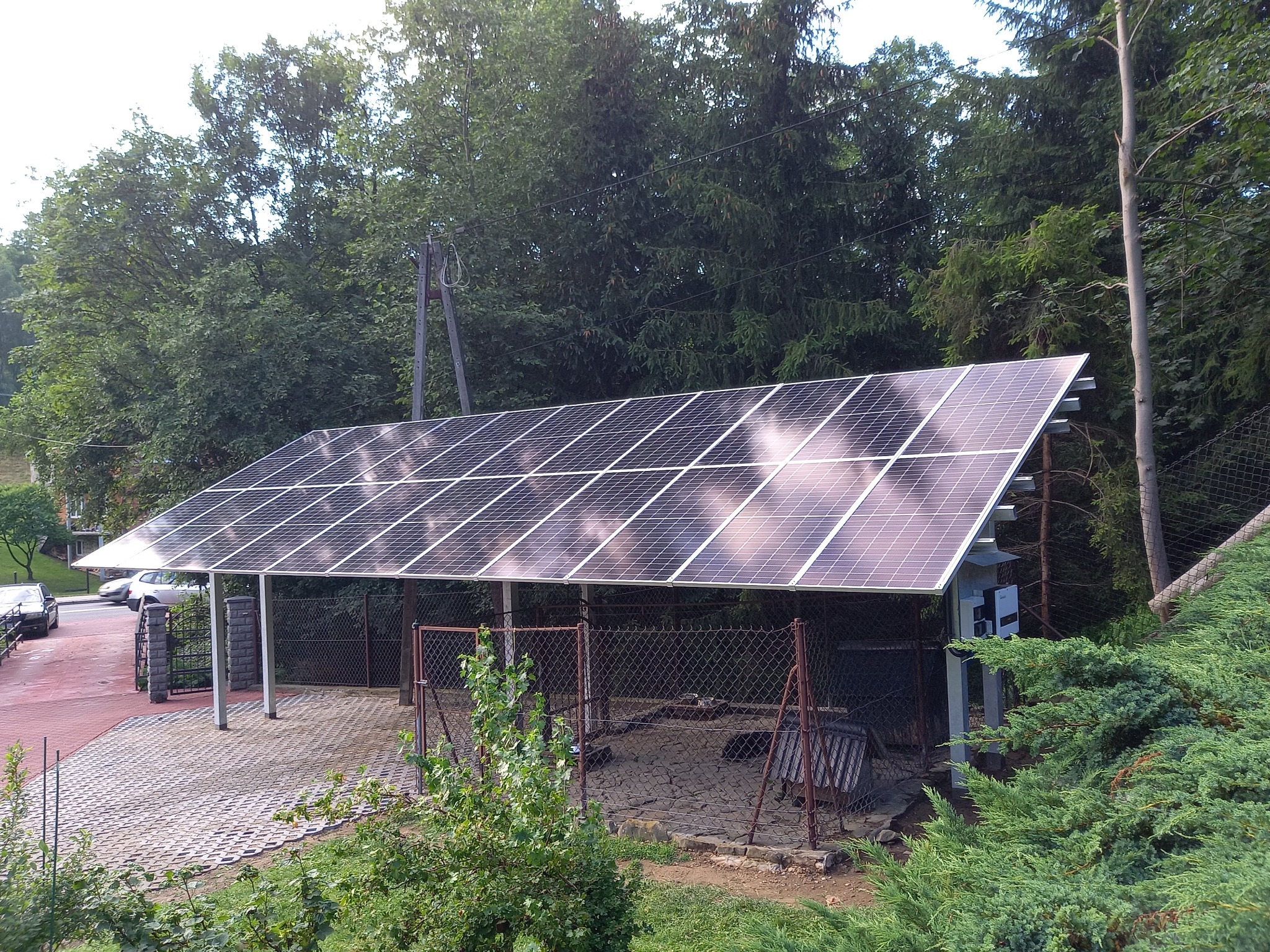 Instalacja fotowoltaiczna na wiacie samochodowej z widocznym inwerterem, otoczona zielenią i drzewami, w tle słup energetyczny.