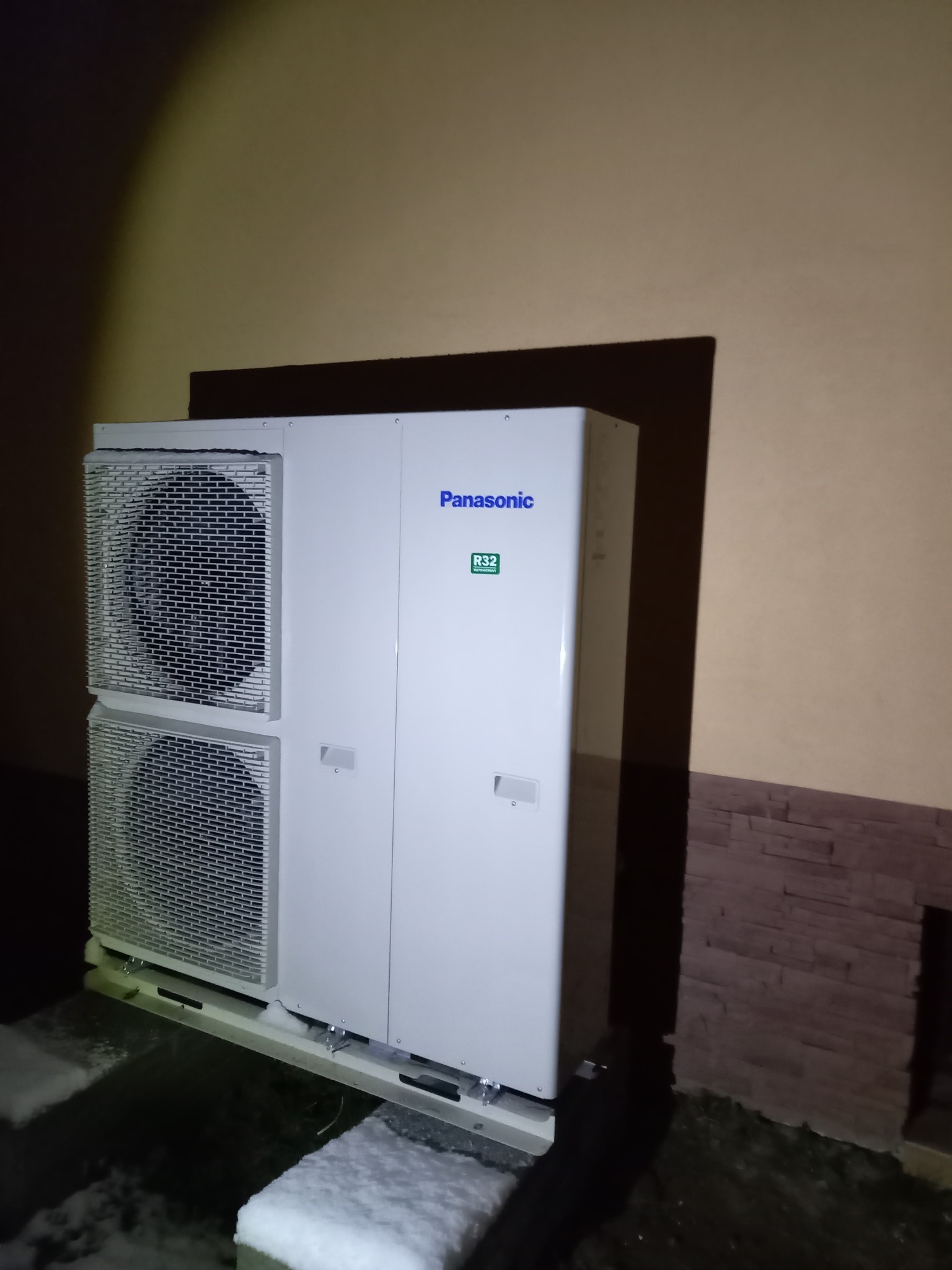 Biała, dwuczęściowa jednostka zewnętrzna z logo Panasonic i oznaczeniem R32, zamontowana przy ścianie budynku z elementami dekoracyjnymi, w zimowych warunkach.
