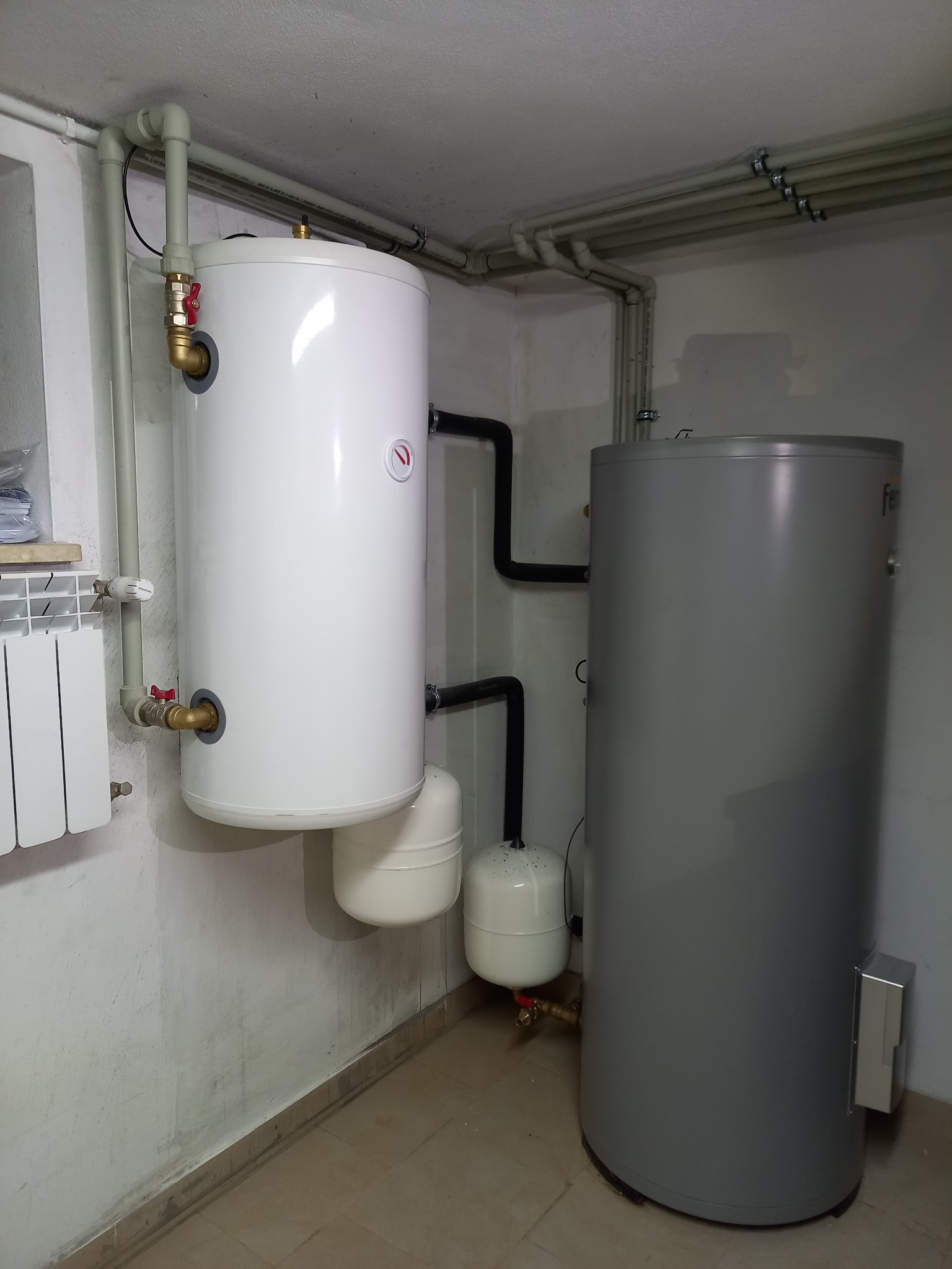 Dwa zbiorniki, biały i szary, połączone czarnymi rurami w piwnicy, z widocznymi zaworami i rurami zasilającymi, obok grzejnik żeberkowy.