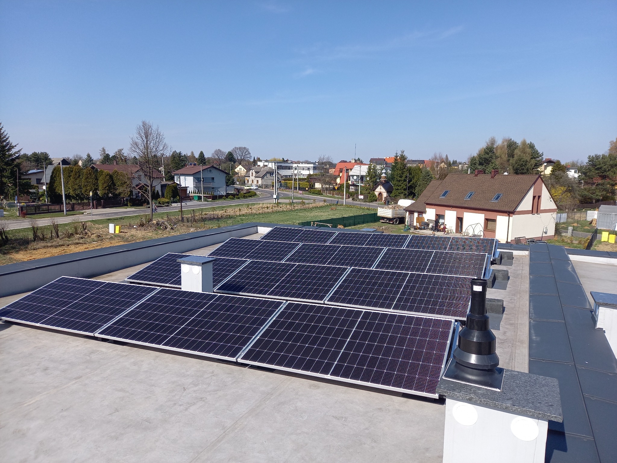 Instalacja fotowoltaiczna na płaskim dachu budynku mieszkalnego, widok z góry na panele w regularnym układzie, komin wentylacyjny z czarnym kołnierzem w prawym dolnym rogu, w tle zabudowa...