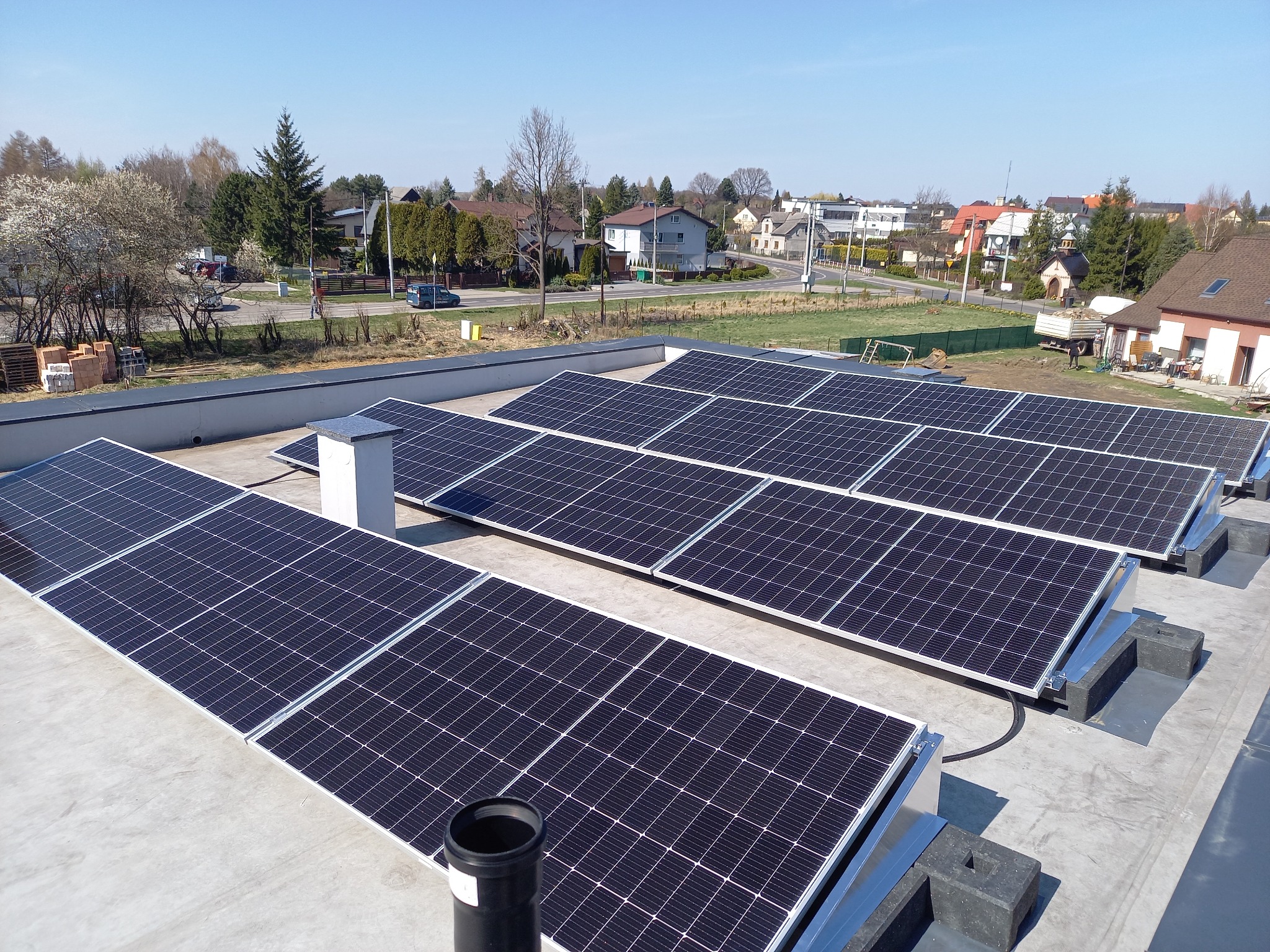 Rząd paneli słonecznych na płaskim dachu budynku mieszkalnego; widok na okoliczne domy i drzewa w słoneczny dzień.