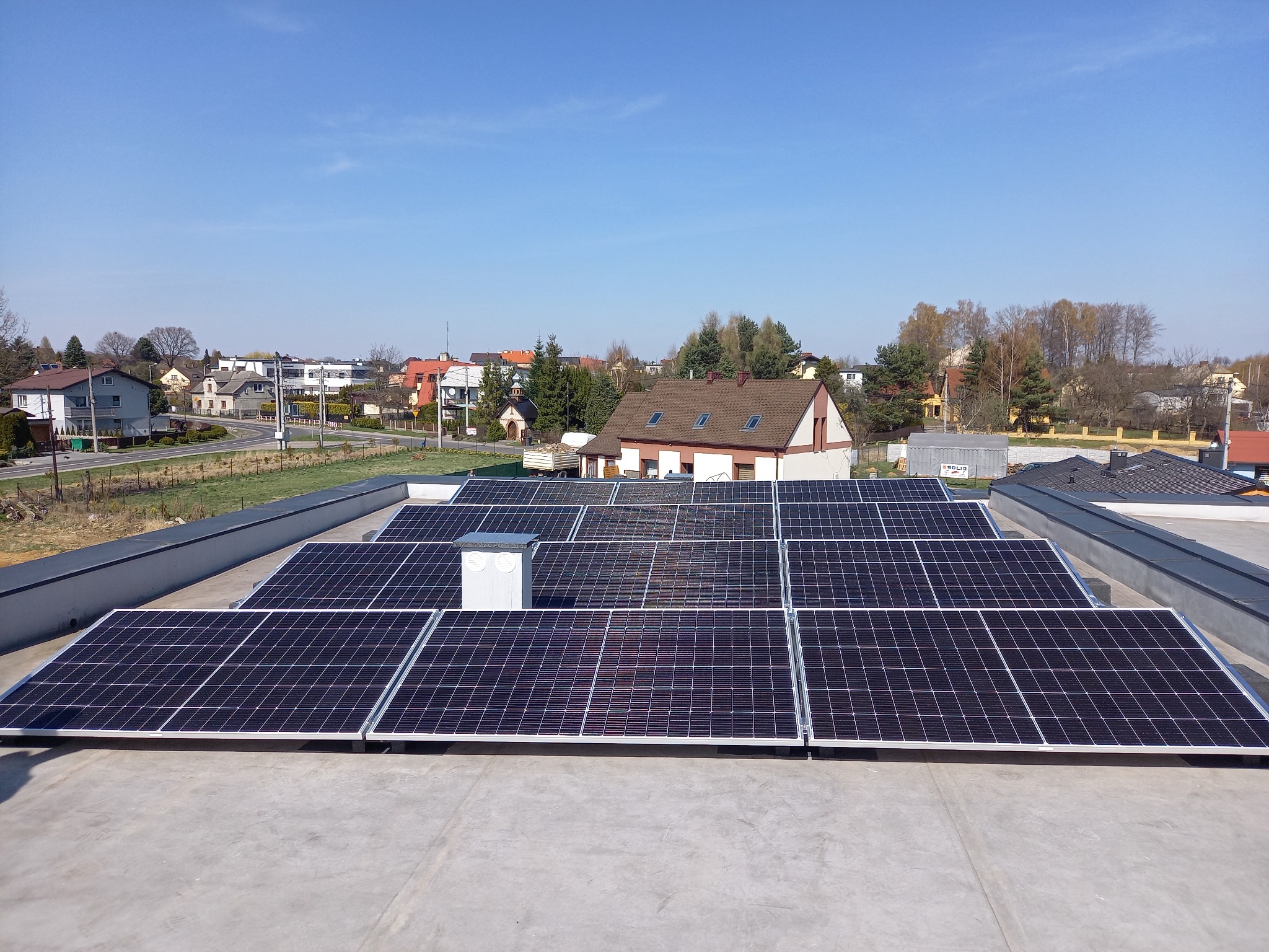 Rząd paneli słonecznych na płaskim dachu budynku, widok z góry w słoneczny dzień, w tle zabudowa jednorodzinna.