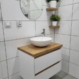 Nowoczesna łazienka z białą umywalką nablatową na szafce z drewnianym blatem, okrągłe lustro i dekoracyjne rośliny na drewnianych półkach na tle białych płytek.