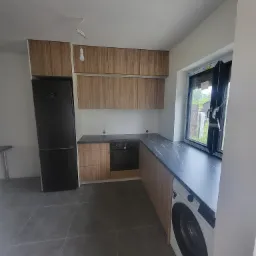 Nowoczesna aranżacja kuchni w Iławie z drewnianymi frontami szafek, czarnym blatem i sprzętem AGD, w tym pralką pod blatem przy oknie.