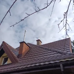 Brązowy dach z metalowej dachówki widziany z dołu, z kominem z rurą i gałęziami drzew na tle pochmurnego nieba.