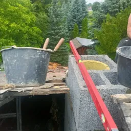 Wznoszenie komina z bloczków betonowych z widoczną izolacją, poziomica przyłożona do ściany komina, wiadro z zaprawą i kielnie na dachu, w tle drzewa.