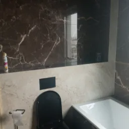 Nowoczesna łazienka z czarną armaturą, marmurowymi płytkami w odcieniach brązu i beżu, białą wanną oraz czarną toaletą z otwartą klapą.