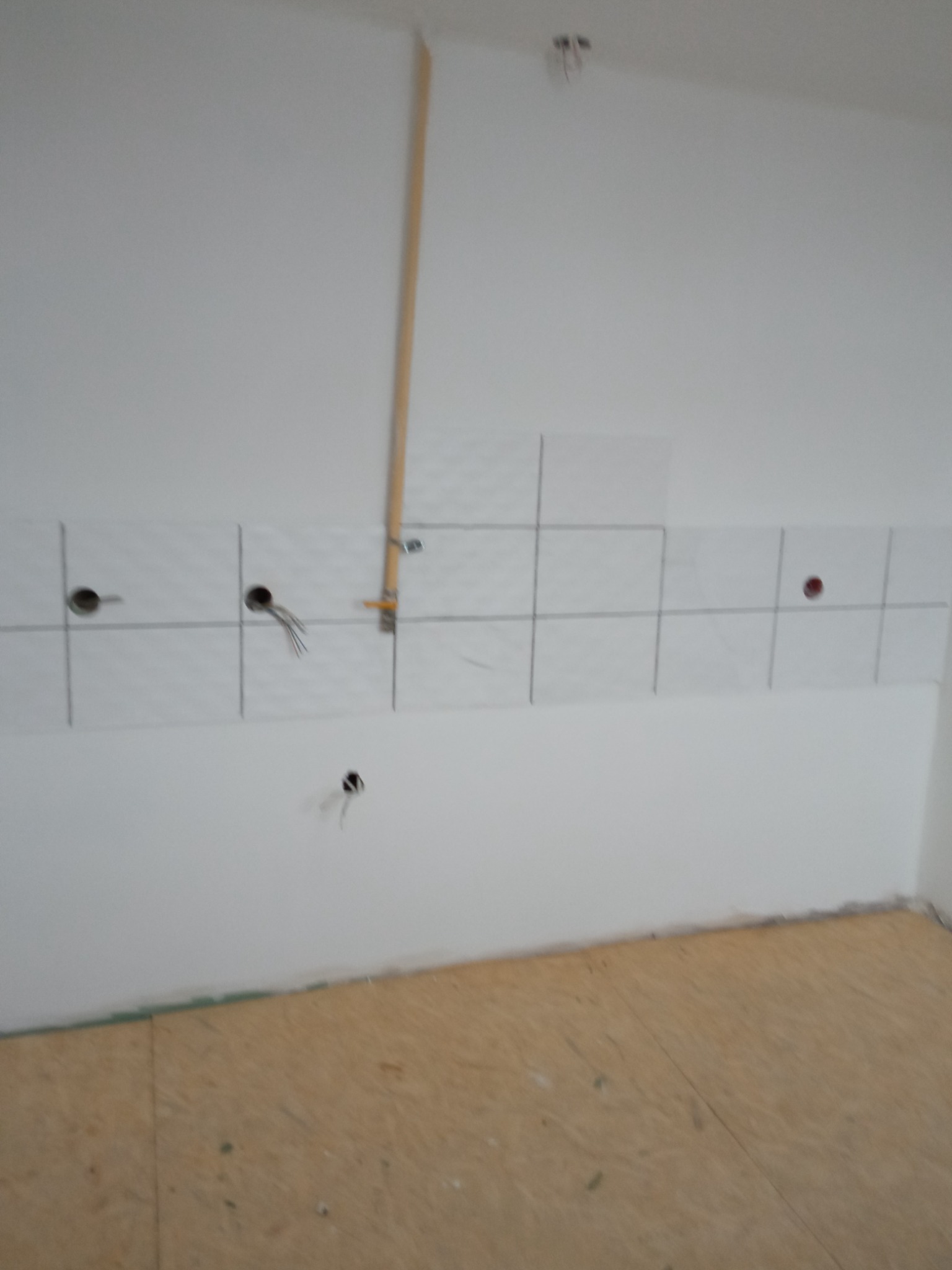 Ściana w trakcie układania białych, kwadratowych płytek z zaznaczonymi liniami fug, widoczne otwory na gniazdka elektryczne, wystające przewody, rurka gazowa i surowa podłoga z płyt OSB.