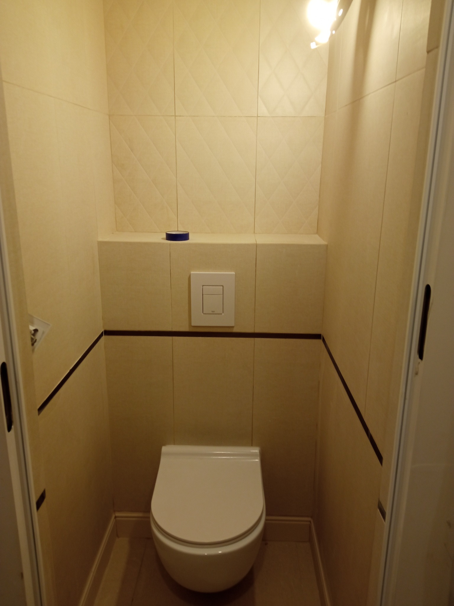 Mała toaleta w beżowych płytkach z dekoracyjnym wzorem rombów na górnej części ścian, z widocznym fragmentem drzwi i niebieskim akcentem dekoracyjnym na półce.