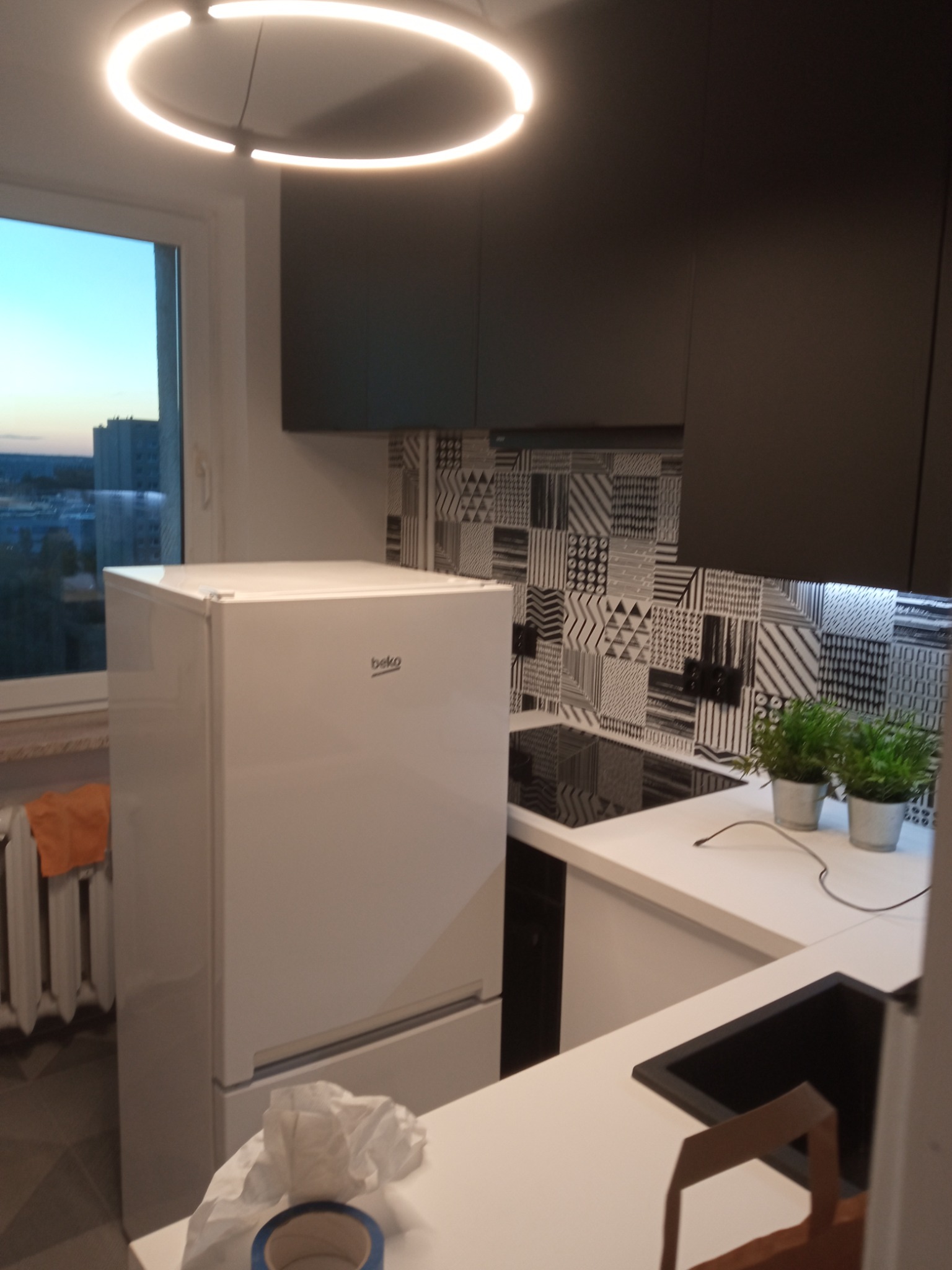 Nowoczesna kuchnia z czarnymi szafkami, wzorzystą mozaiką na ścianie, białym blatem, zlewem i lodówką Beko, oświetlona okrągłą lampą sufitową i listwą LED pod szafkami wiszącymi.