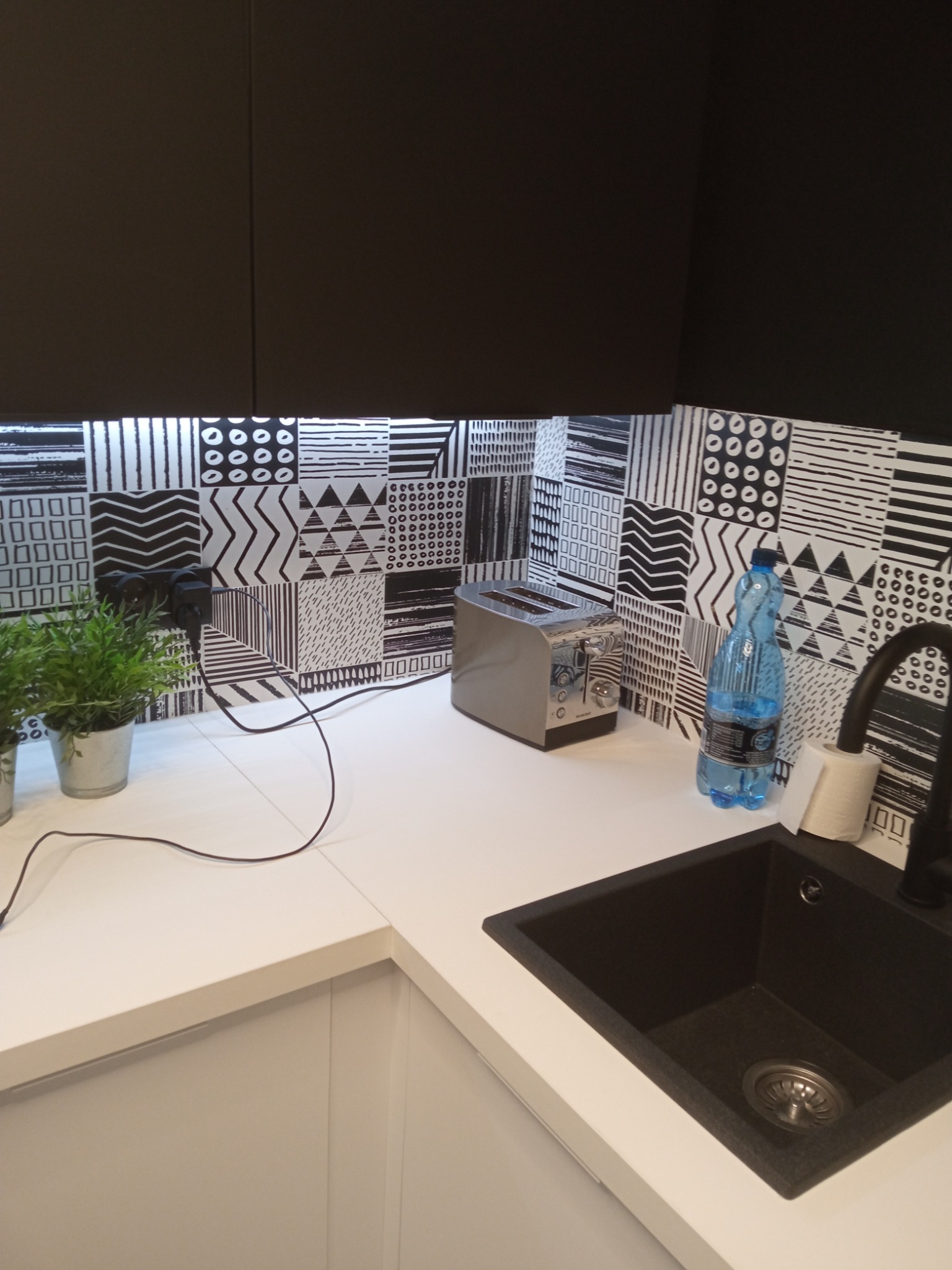 Nowoczesna kuchnia z czarnymi szafkami, białą zabudową, czarnym zlewem i baterią, oraz wzorzystą mozaiką na ścianie nad blatem, uzupełniona o toster, butelkę wody i doniczkę z rośliną.